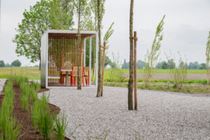 Offener Pavillon mit Holzlamellen, Tisch und roten Stühlen in einer gepflegten Außenanlage mit jungen Bäumen, Kiesweg und Wiesen im Hintergrund.